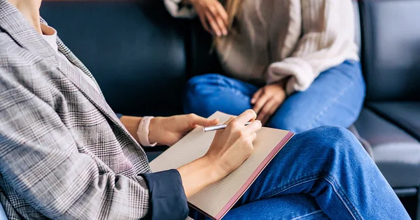 A counselor or behavioral health professional takes notes during a session, focusing on active listening and documentation, with a client partially visible.