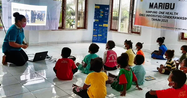 Janet Matani with a face mask gives a presentation to a group of children seated on the floor, with a projected image on a screen and a banner in the background.