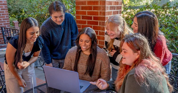 A group of six college students smiling and looking at a laptop together outdoors.