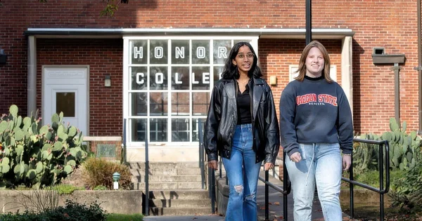 Two female students walking outside the entrance of a red brick building with 'HONORS COLLEGE' printed on the large windows.