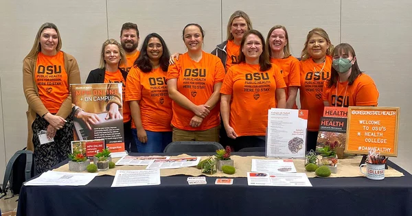 A large, enthusiastic team from the College of Health posing together at an event booth with promotional materials for graduate programs.