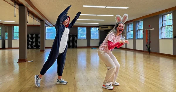 Two people in a brightly lit gymnasium posing playfully in costumes: one dressed as a shark with arms raised, and the other as a rabbit with bunny ears and a crouched posture.