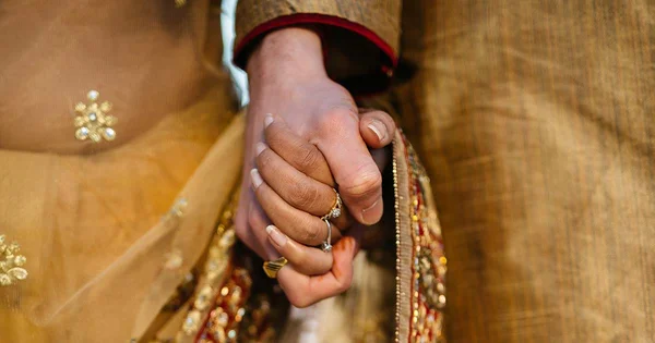  Close-up of a man and woman holding hands, symbolizing love and union. Both are wearing traditional Indian or South Asian wedding attire in rich gold and rust tones.