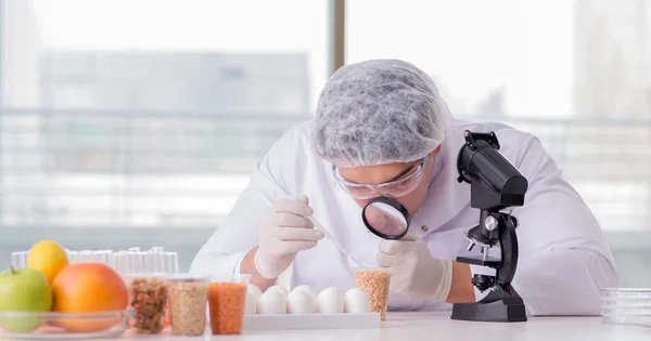 A food science researcher in a lab coat, hairnet, and safety glasses uses a magnifying glass to closely inspect a sample of grain or cereal, with eggs and fruit nearby