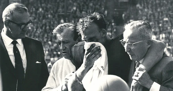 Black and white photograph of an emotional exchange on a football sideline, where coaches comfort a player whose face is covered by a towel.