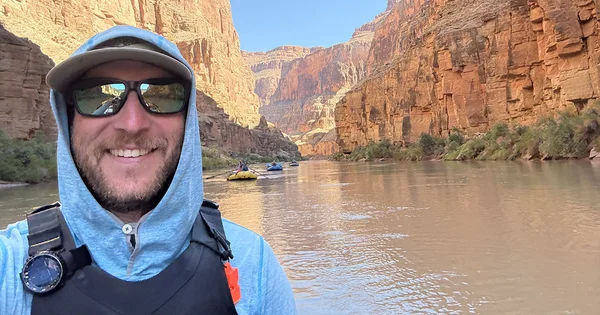 A man wearing a hooded sun shirt, hat and sunglasses smiles while rafting through a canyon on a calm river. Steep orange rock walls rise on both sides and several rafts float in the background.