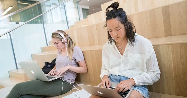 Two female students sit on wide wooden steps in a modern campus building, concentrating on their laptops while wearing earbuds or headphones.