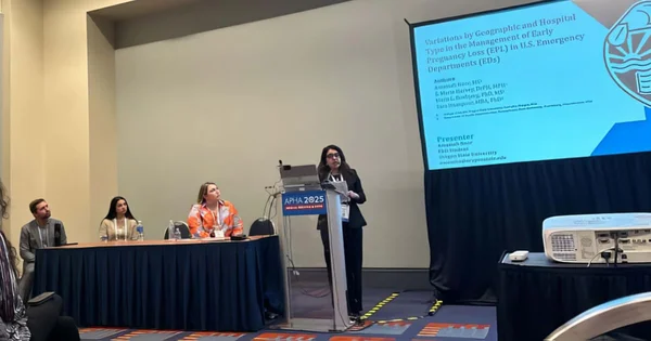 A presenter speaking at a podium during the APHA 2025 conference, with her research title displayed on a screen.
