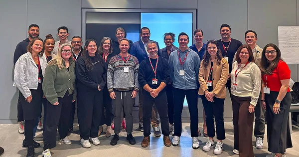 A large group of approximately twenty colleagues or professionals, wearing lanyards and casual attire, stands together for a group photo in front of a bright screen