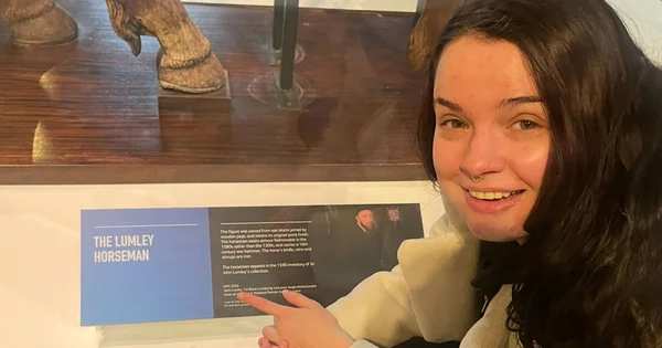 Lizzie Lumley smiles and points to a museum display labeled “The Lumley Horseman” while standing beside a glass case holding carved wooden figures.
