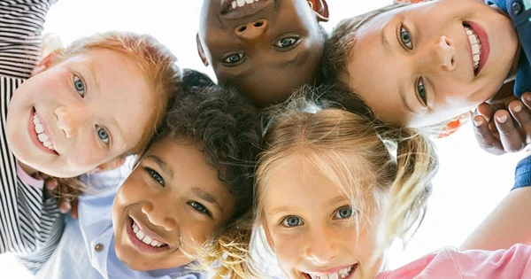 A close-up, low-angle shot of five diverse, smiling children looking down at the camera.