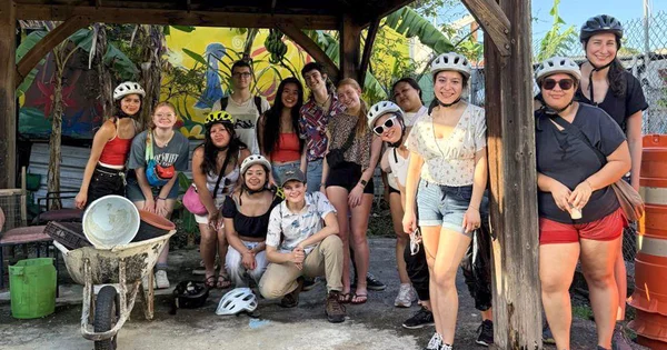 A diverse group of 14 smiling university students wearing helmets pose under a wooden outdoor shelter in a tropical or communal farming setting.