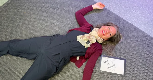 A woman named Dee collapses dramatically on the floor, celebrating an achievement after a long project, as indicated by the congratulatory sign.