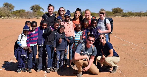 A university team and local children posing together in a sunny, arid landscape, symbolizing a global service project or study abroad initiative.