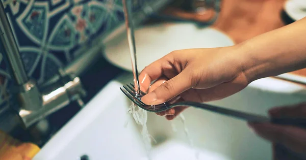 A close-up of a hand rinsing a metal fork under running water in a kitchen sink.
