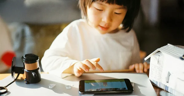 A young girl with bangs, wearing a white shirt, leans over a desk to touch a horizontally held smartphone screen. Sunlight shines on a small, clipped black microscope and a paper model of a house.