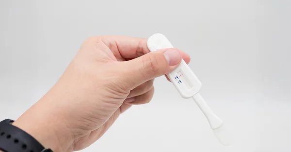 A close-up of a hand holding a white rapid diagnostic test against a plain light gray background.
