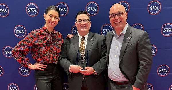 Three smiling individuals pose in front of a blue backdrop patterned with the Student Veterans of America (SVA) logo. The person in the center holds a glass award.