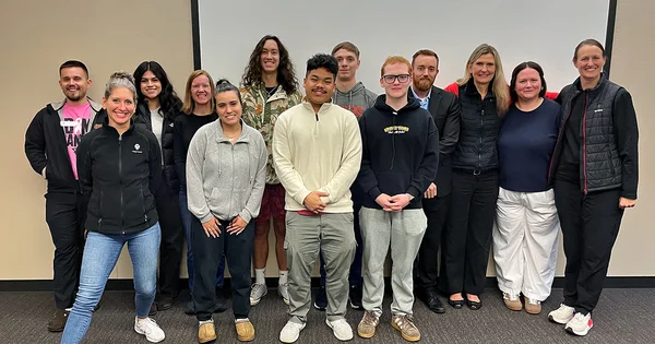 A diverse group of 14 smiling students and faculty members stand in two rows against a neutral wall in a professional meeting room setting.