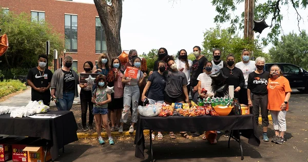 A large group of approximately 20 people, most wearing face masks, pose behind a table filled with snacks and drinks at an outdoor community event on a sunny day.