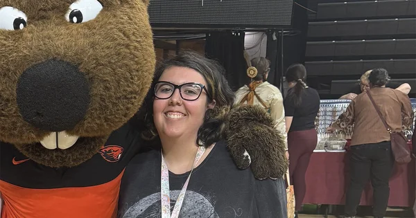 Attendee at an indoor event posing with a beaver mascot, with booths and other people in the background.