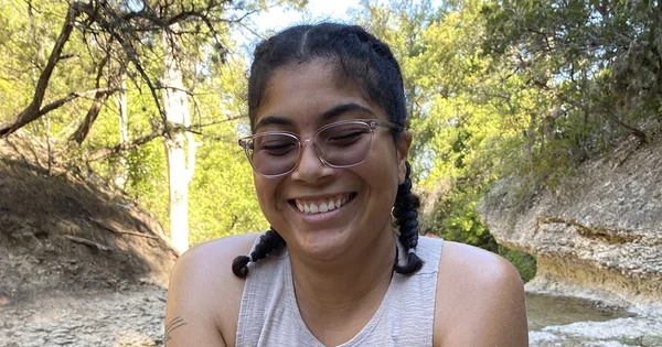 Kylee Sharp Gonzalez smiling in a natural creek setting with trees and rocky banks in the background.