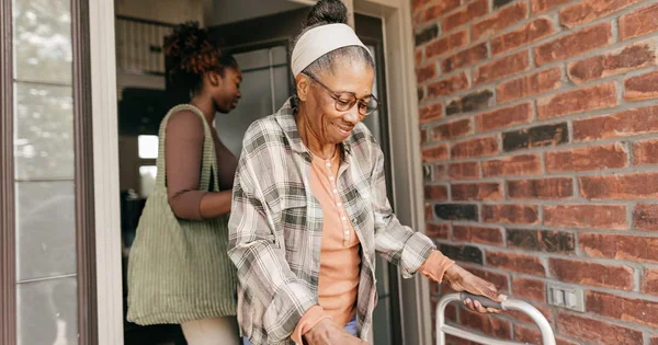 A caregiver helping an elderly woman with a walker leave her home. Emphasizes independence and support.