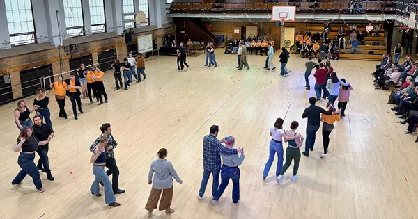 Groups of couples line dancing in a gymnasium with a wood floor and audience seating in the background.