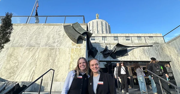 Two smiling women stand in front of the marble facade and rotunda of the Oregon State Capitol under a clear blue sky.