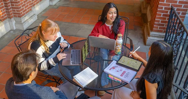 Diverse college students engaged in collaborative group study at an outdoor table using digital tools and printed materials.