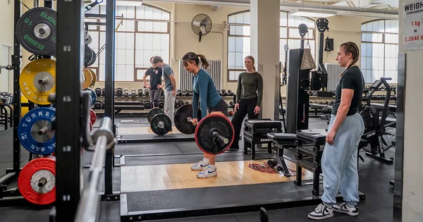 Woman in blue shirt deadlifting a barbell with red plates on a wooden platform, observed by another woman in a busy gym setting.