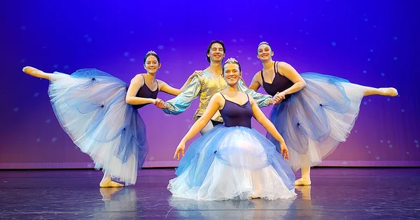 Four ballet dancers in blue tutus and a silver tunic perform a choreographed pose against a purple and blue starry stage backdrop.