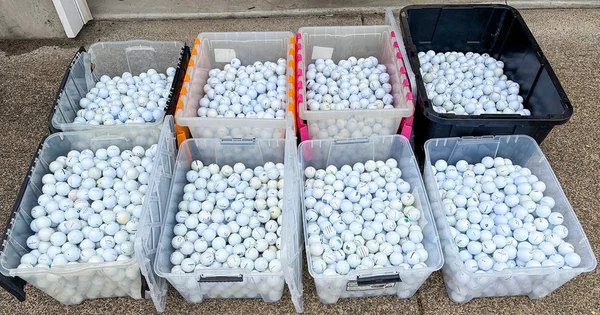 Seven large plastic bins filled with hundreds of white golf balls, arranged in two rows on a paved surface.
