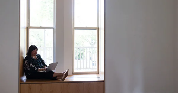 A woman sits in a window nook using a laptop, silhouetted by bright daylight from two large windows.