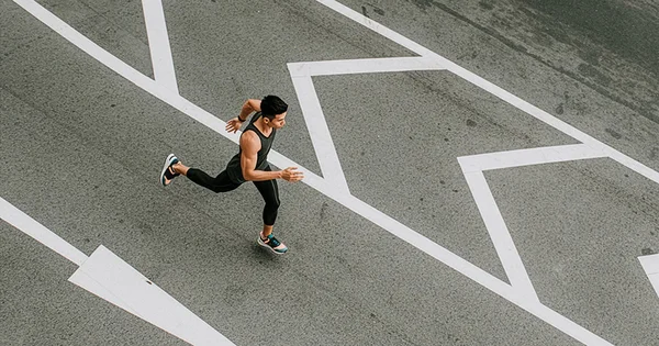 High-angle view of a man in black athletic wear running across a gray asphalt road with bold white geometric markings.