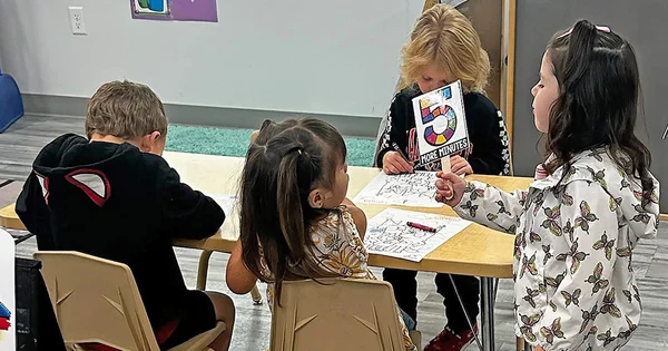 Four young children sit at a table coloring; one girl holds up a sign that reads '5 MORE MINUTES' featuring a colorful number five.