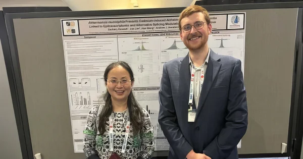 Two researchers, a man in a suit and a woman in a patterned shirt, smile while standing in front of a scientific research poster at a conference.