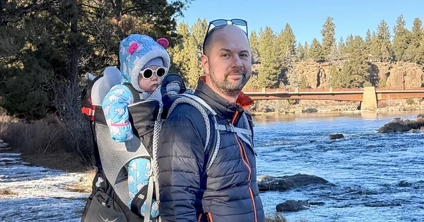 Man wearing a puffer jacket carrying a baby in a blue snowsuit and sunglasses in a backpack carrier next to a flowing river.