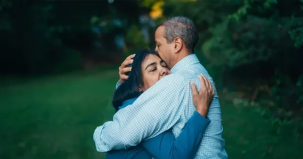 Man with graying hair gently kisses the forehead of a woman with dark hair, eyes closed, in a tender embrace outdoors.