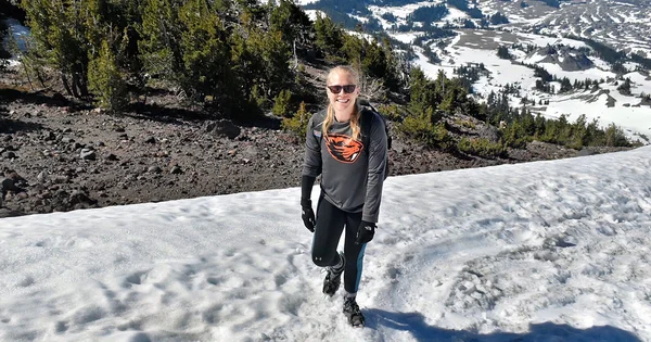 Hiker wearing an Oregon State Beavers shirt smiles while navigating a snowy trail on a high mountain peak overlooking a scenic alpine valley.