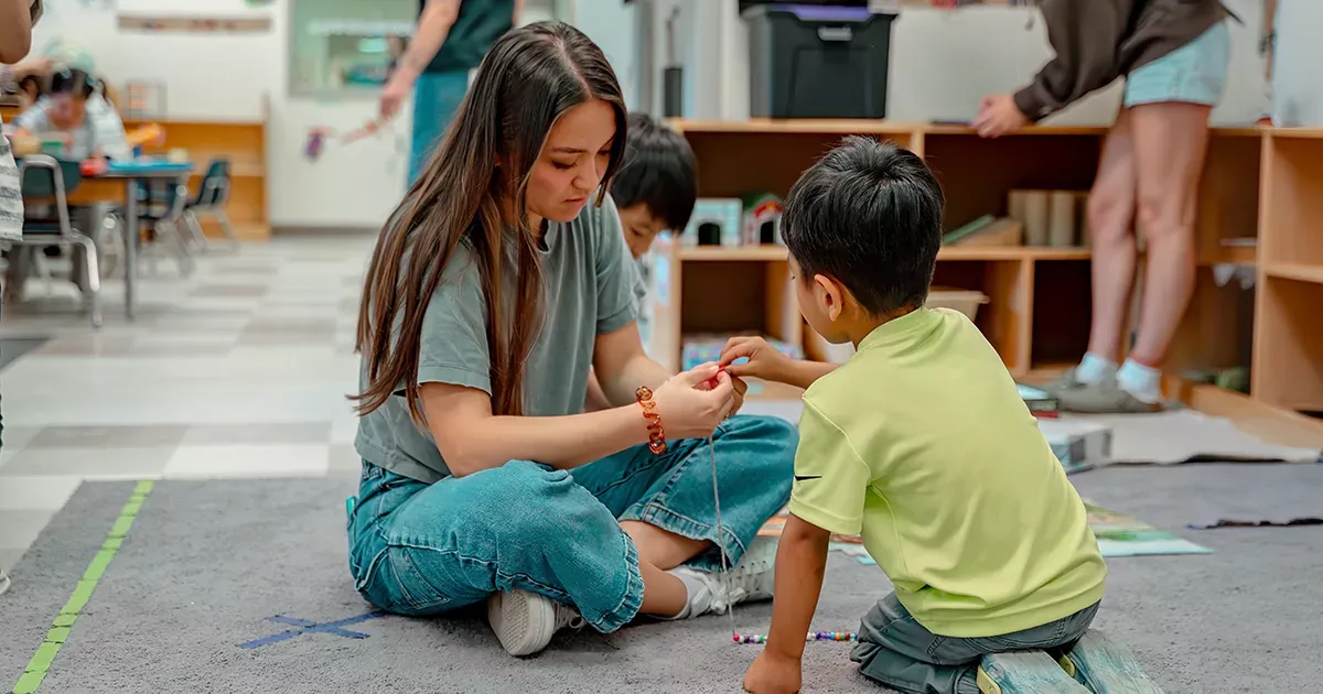 College student and preschool child sitting on the floor of a classroom, engaged in a hands-on learning activity.