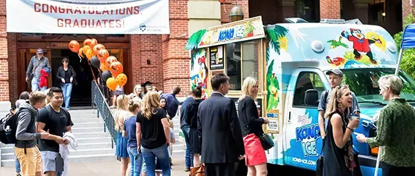  A group of people gathered outside a building, with a banner that says "CONGRATULATIONS, GRADUATES!" and a colorful ice cream truck in the background.