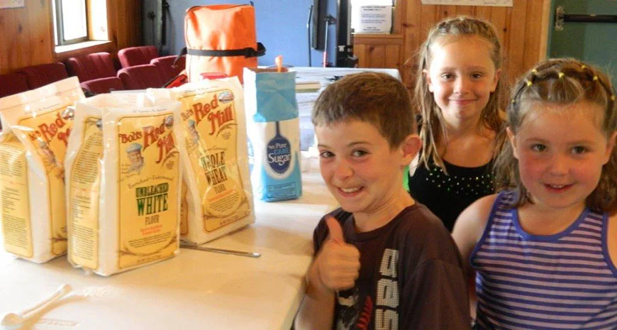  Three children posing in a room with various household items and food items in the background.