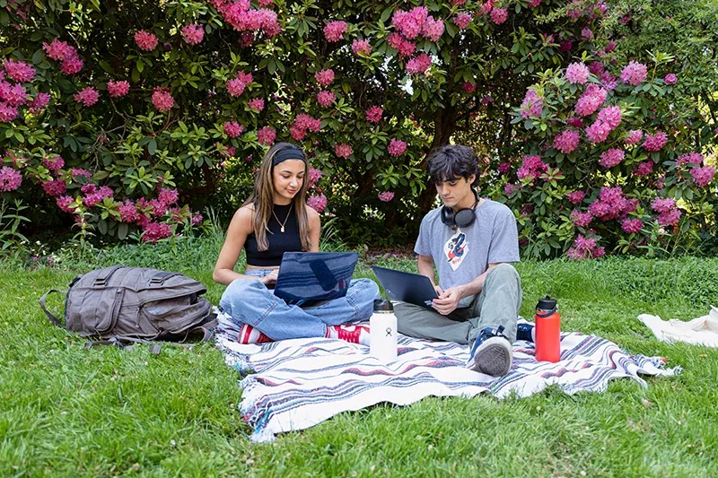 Two people sitting on a blanket on grass on campus with blooming flowers.