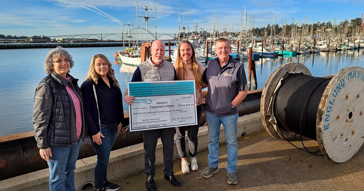 A group of five people standing in front of a marina with boats and a bridge in the background, holding a large check.