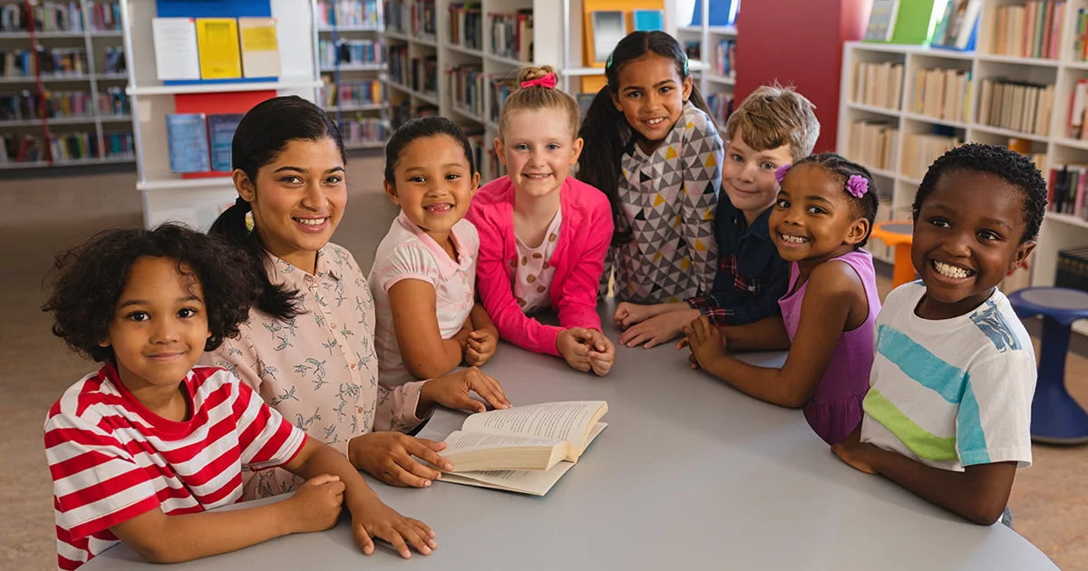 Group of diverse children sitting together at a table in a library, with an open book in front of them.