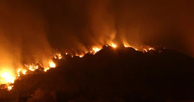 Nighttime wildfire burning along the ridge of a dark mountain, with intense orange and yellow flames spreading across the landscape.