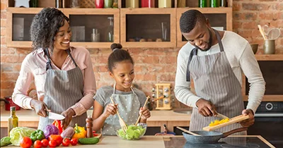 Family of three preparing food together in a kitchen, with fresh vegetables on the counter.