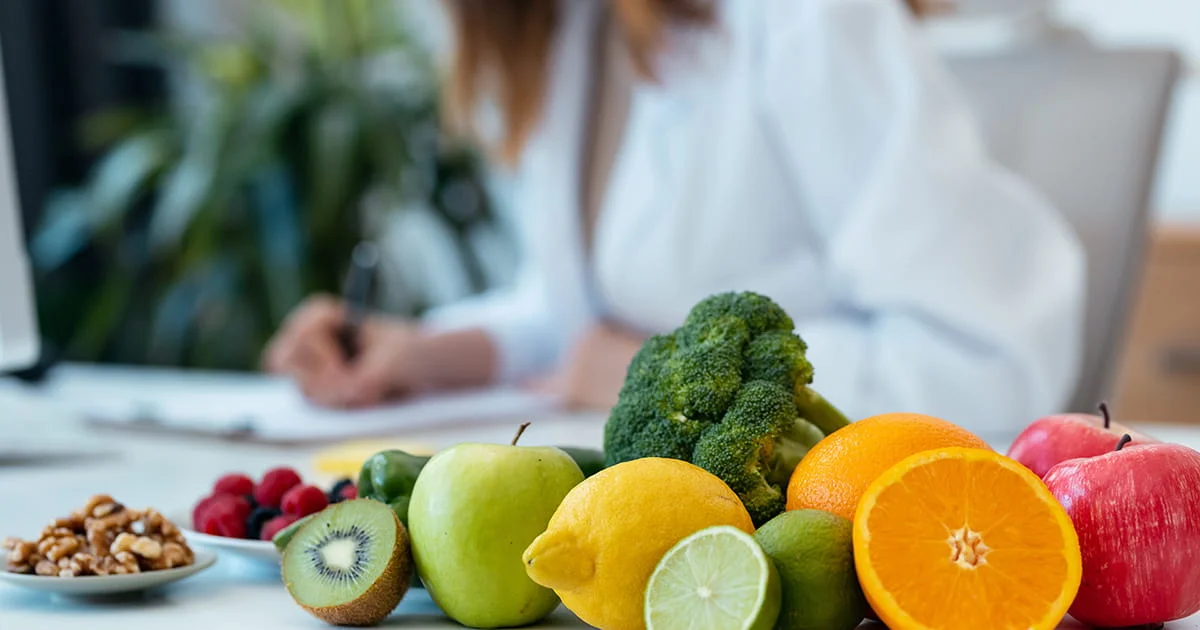 Assorted fresh fruits and vegetables including broccoli, oranges, apples, kiwi, lemons, and limes on a table with a blurred background of someone working.