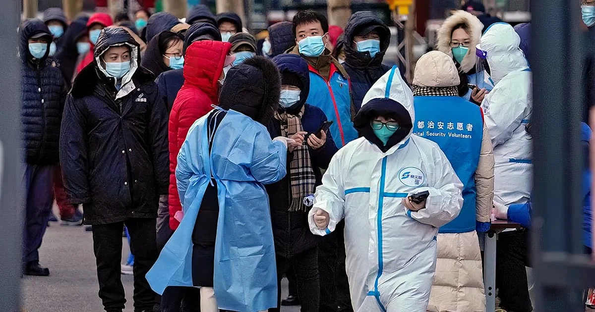 Crowd of people wearing winter coats and medical masks, with some in protective medical gear, standing on a street.